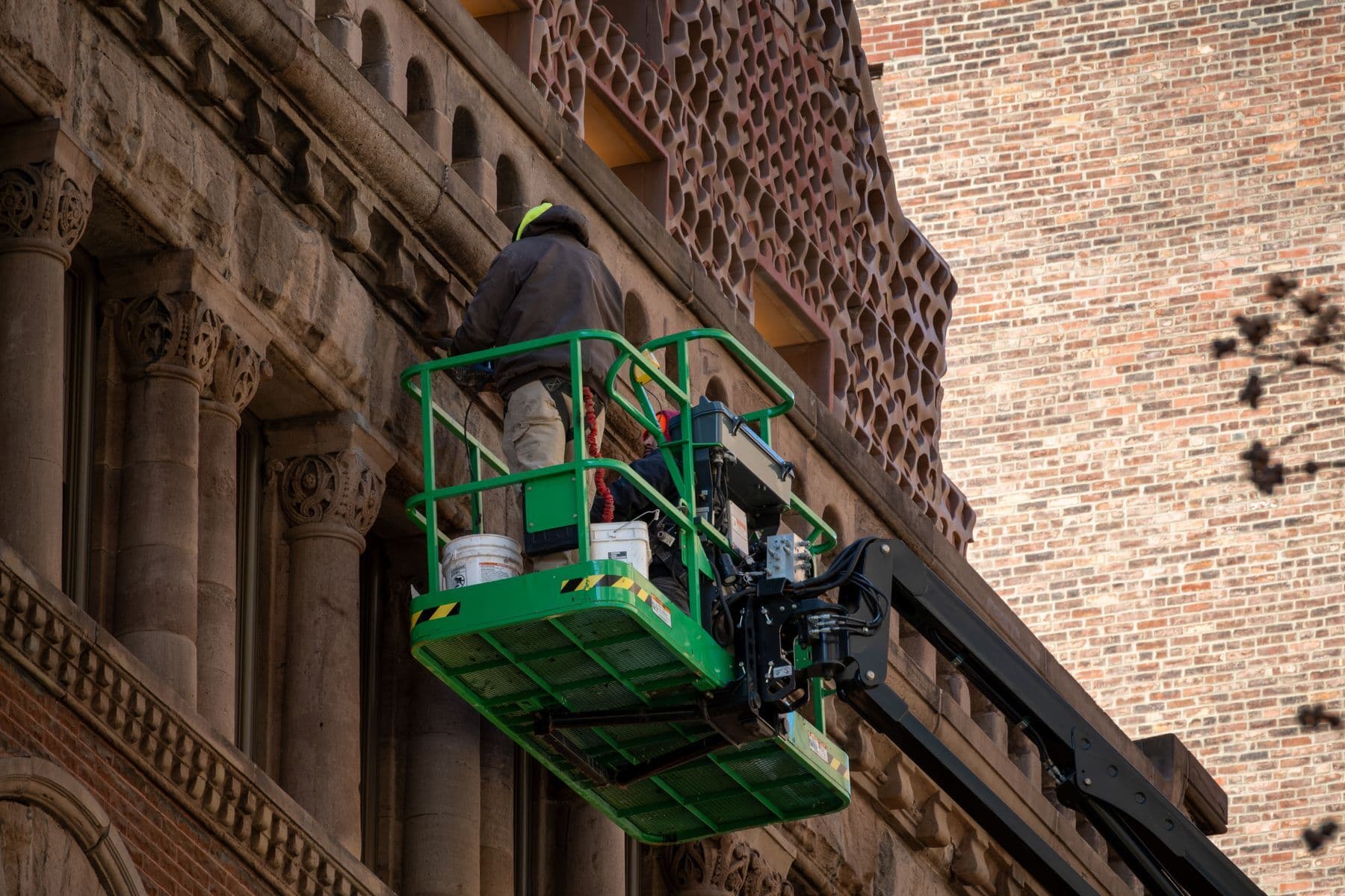 A worker in a green boom-lift basket coating the carved stone and brick facade of an old masonry building.
